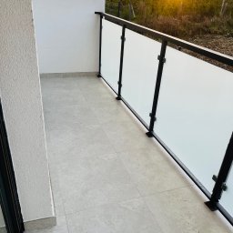 ProEfekt - Wykończony balkon z jasnymi płytkami podłogowymi i nowoczesną balustradą z czarną ramą i mlecznym szkłem, widok na zachodzące słońce za drzewami.
