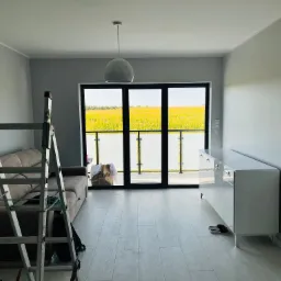 Jasne wnętrze pokoju po odświeżeniu, z drabiną malarską, szarymi ścianami, białymi meblami i dużym oknem balkonowym z widokiem na pole.