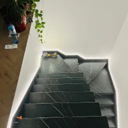 Schody wyłożone czarnym gresem z białymi żyłami, podświetlone listwą LED, z narzędziem do cięcia płytek na górnym stopniu, widok z góry.