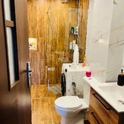 Nowoczesna łazienka z drewnopodobnymi płytkami, kabiną prysznicową typu walk-in i pralką. Umywalka z drewnianą szafką, WC kompakt. Jasne, ciepłe oświetlenie.