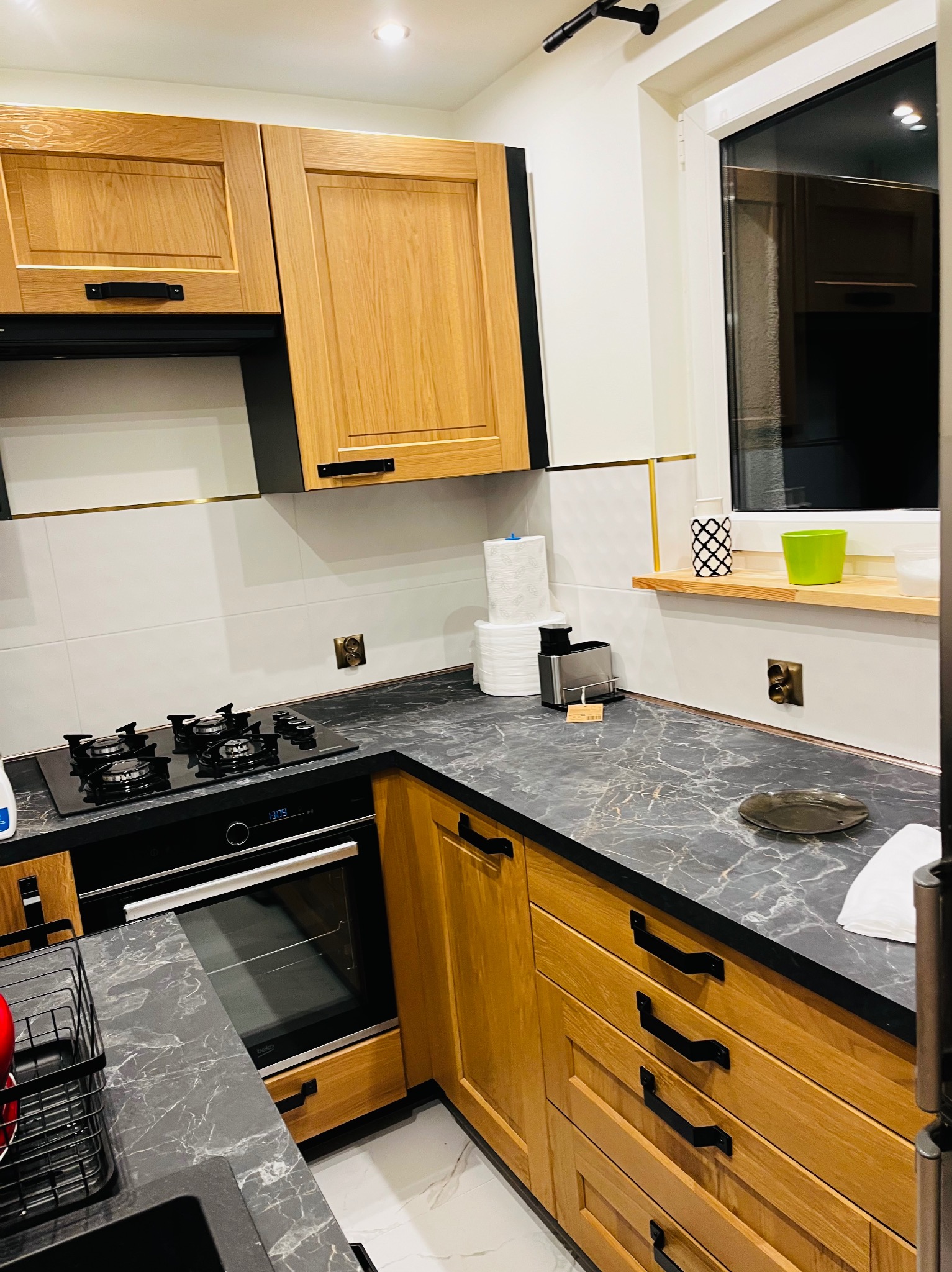 Nowoczesna kuchnia z drewnianymi szafkami, czarnym blatem imitującym marmur, płytą gazową i piekarnikiem w zabudowie, wykończona białymi płytkami z delikatnym złotym akcentem.