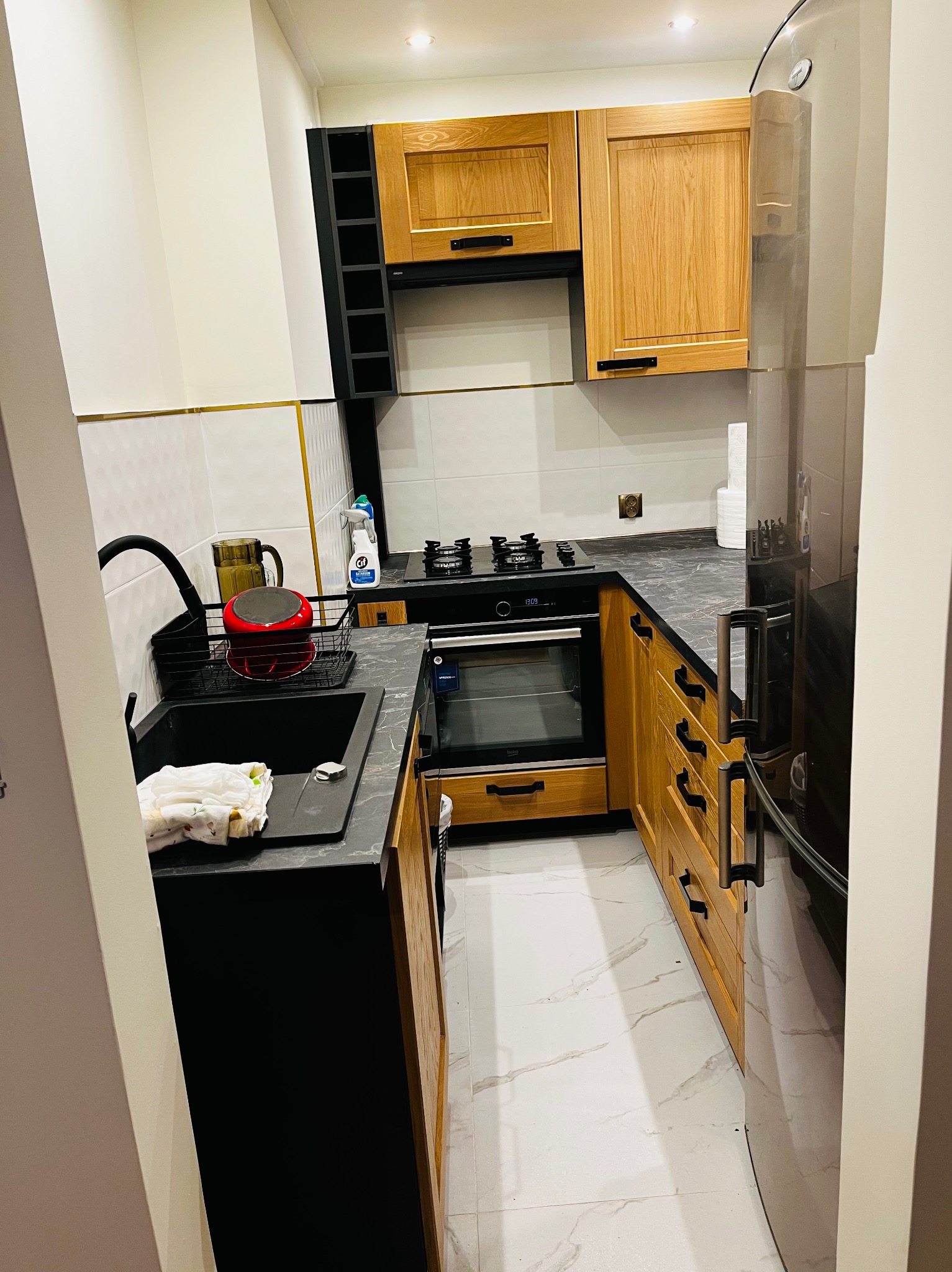 Nowoczesna, kompaktowa kuchnia w odcieniach bieli, drewna i czerni, z marmurową podłogą i blatem, wyposażona w piekarnik, płytę grzewczą, zlewozmywak i lodówkę, z akcesoriami kuchennymi na wierzchu.