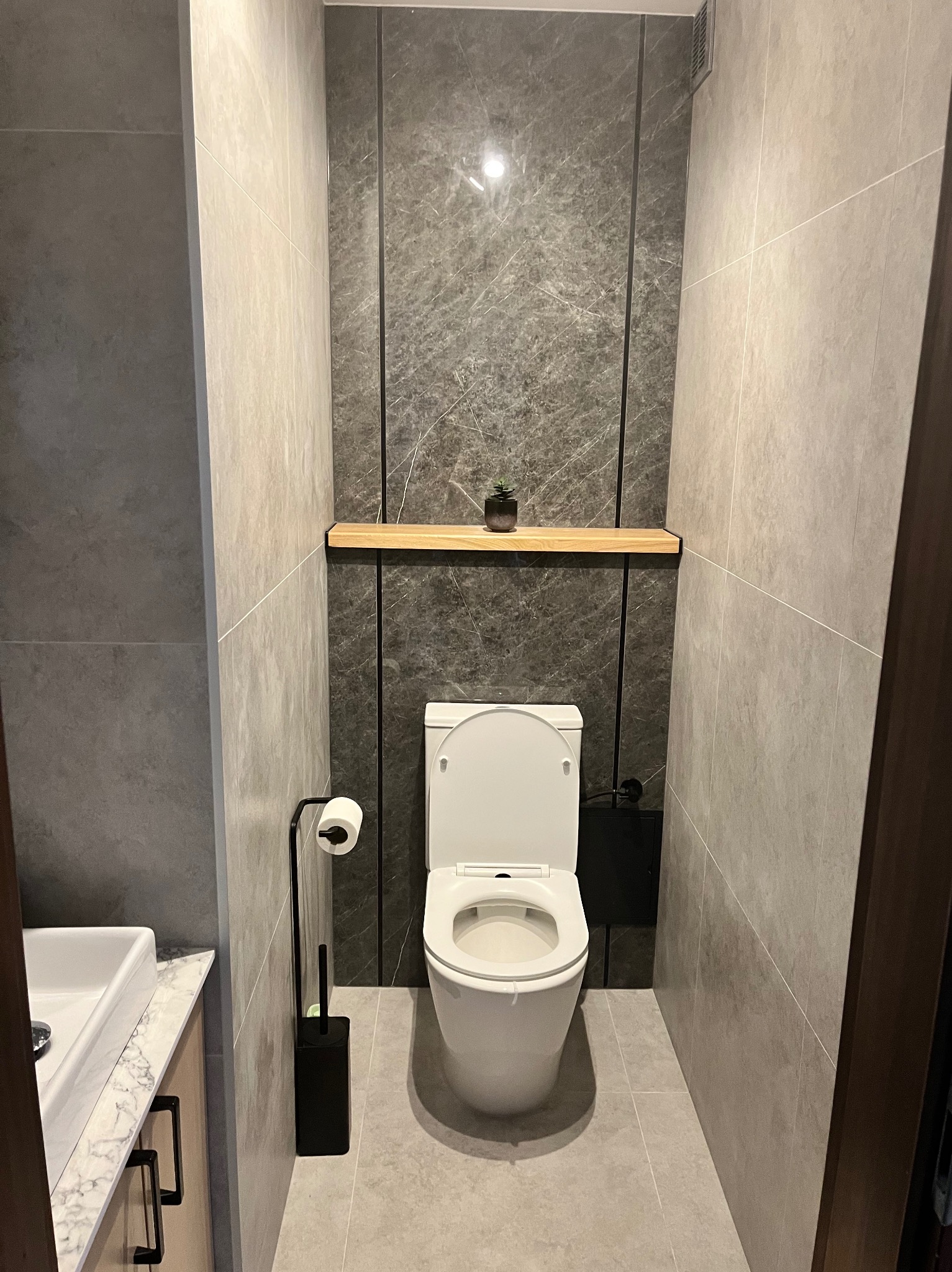 Nowoczesna toaleta w wąskiej łazience wykończonej szarymi płytkami, z drewnianą półką nad ubikacją i małą roślinką doniczkową.
