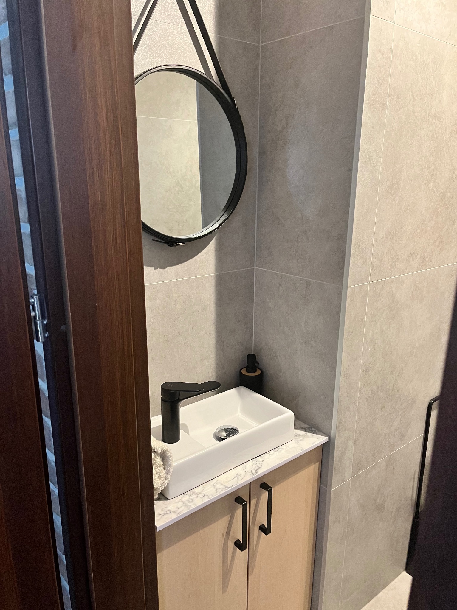 Umywalka nablatowa z czarną armaturą, okrągłe lustro na pasku i beżowe płytki w wykończonej łazience.