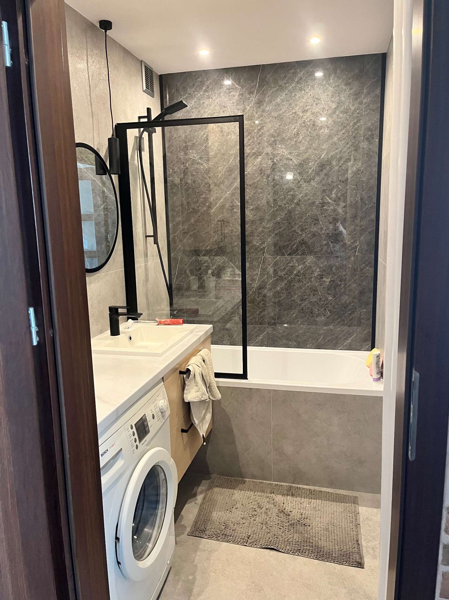 Nowoczesna łazienka z kabiną prysznicową typu walk-in z czarną ramą, umywalką nablatową, pralką Bosch i szarą marmurową okładziną ścienną.