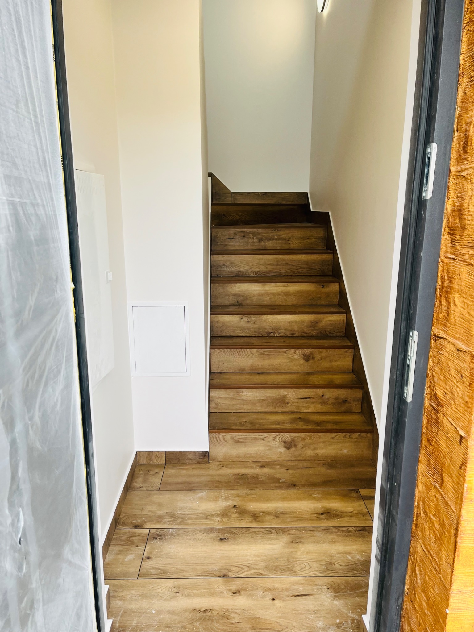 Klatka schodowa wykończona panelami imitującymi drewno, z widoczną dolną częścią schodów i fragmentem podłogi, świeżo po pracach wykończeniowych, z zabezpieczoną folią ościeżnicą drzwi.