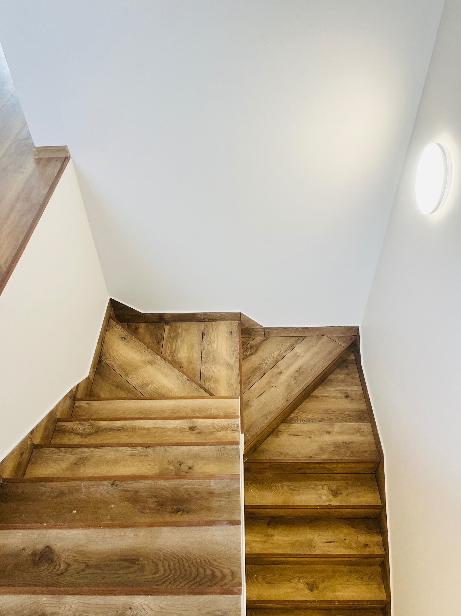 Drewniane schody z panelami ściennymi w jodełkę, oświetlenie sufitowe LED, białe ściany.