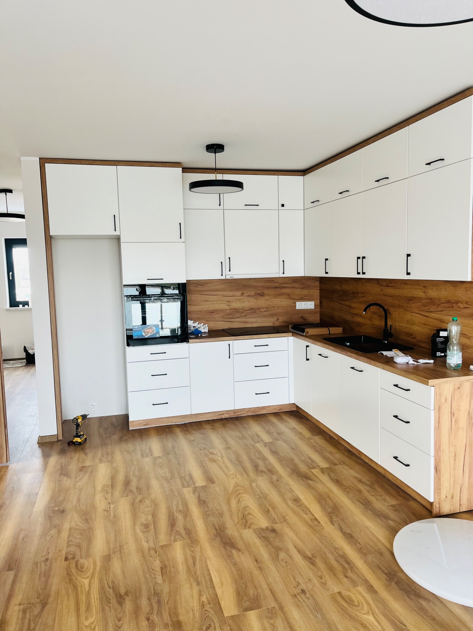 Nowoczesna kuchnia z białymi szafkami, drewnianym blatem i okładziną ścienną, czarną armaturą i lampą sufitową. Na podłodze panele imitujące drewno. W rogu wiertarka akumulatorowa.