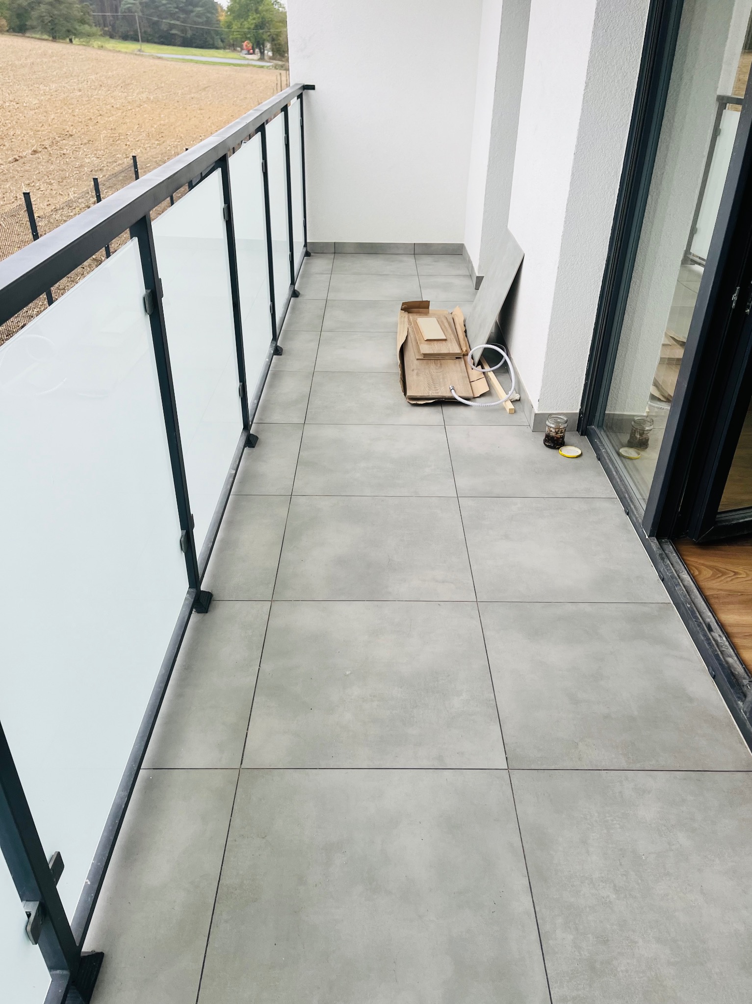 Szare płytki gresowe na podłodze balkonu z przeszkloną balustradą, widoczne niedokończone prace wykończeniowe, karton z panelami i słoik z akcesoriami przy drzwiach balkonowych.