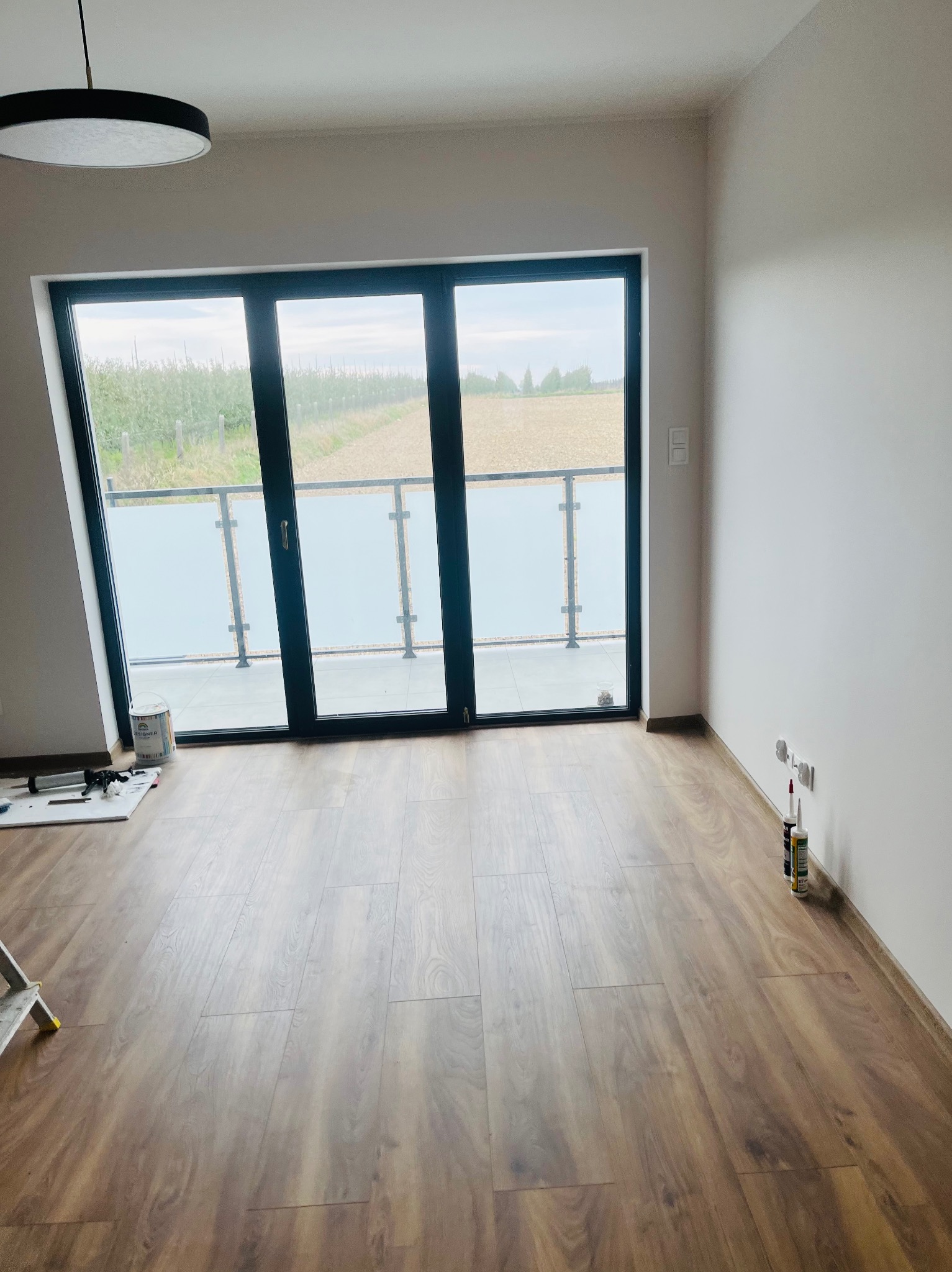 Pusty pokój z drewnianą podłogą i dużym oknem balkonowym z widokiem na sad, przygotowany do prac wykończeniowych, widoczne akcesoria malarskie i drabina.