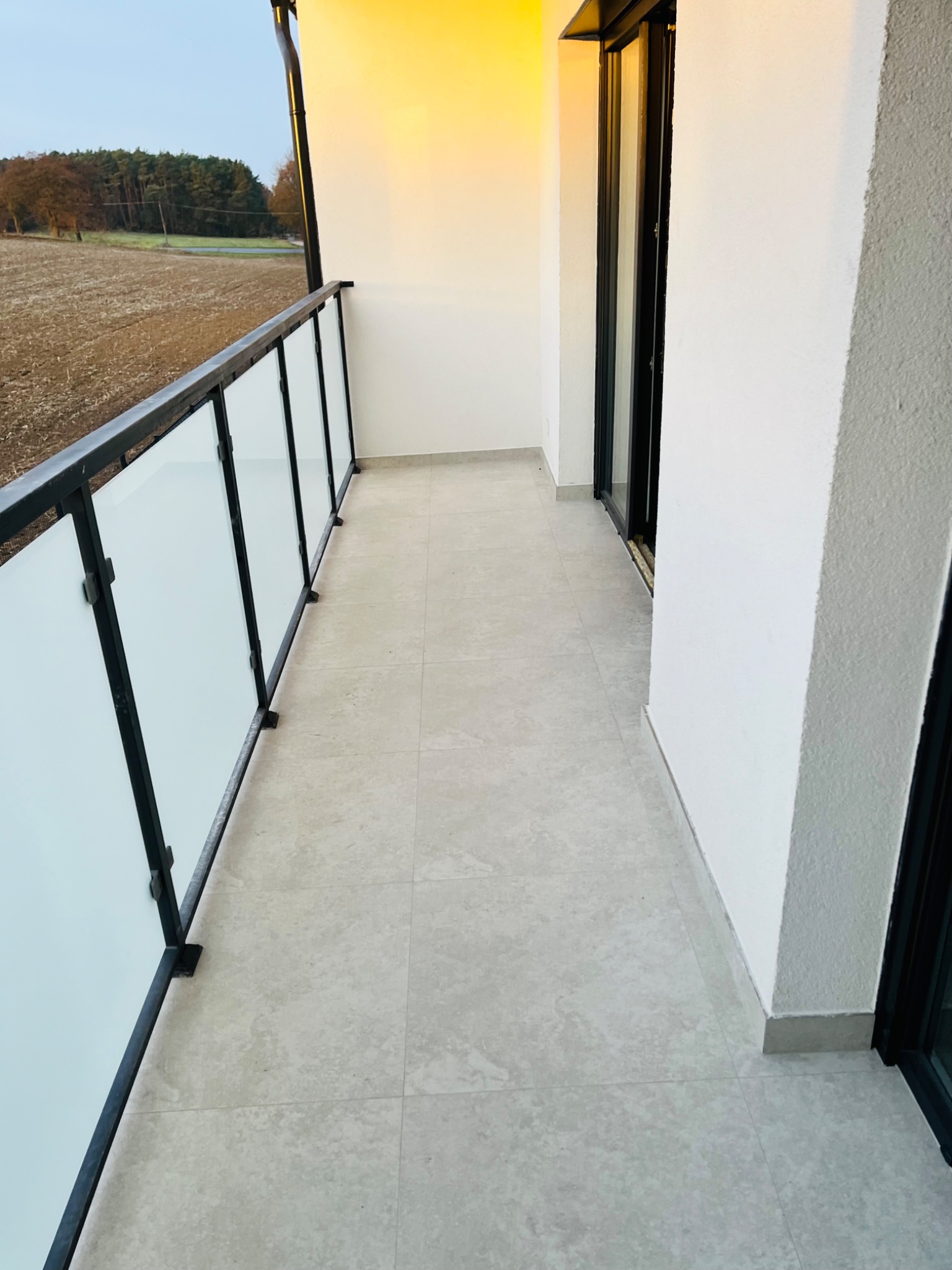 Jasny balkon z szarą, płytkowaną podłogą, przeszkloną balustradą z czarnymi elementami i widokiem na pole z drzewami w oddali.