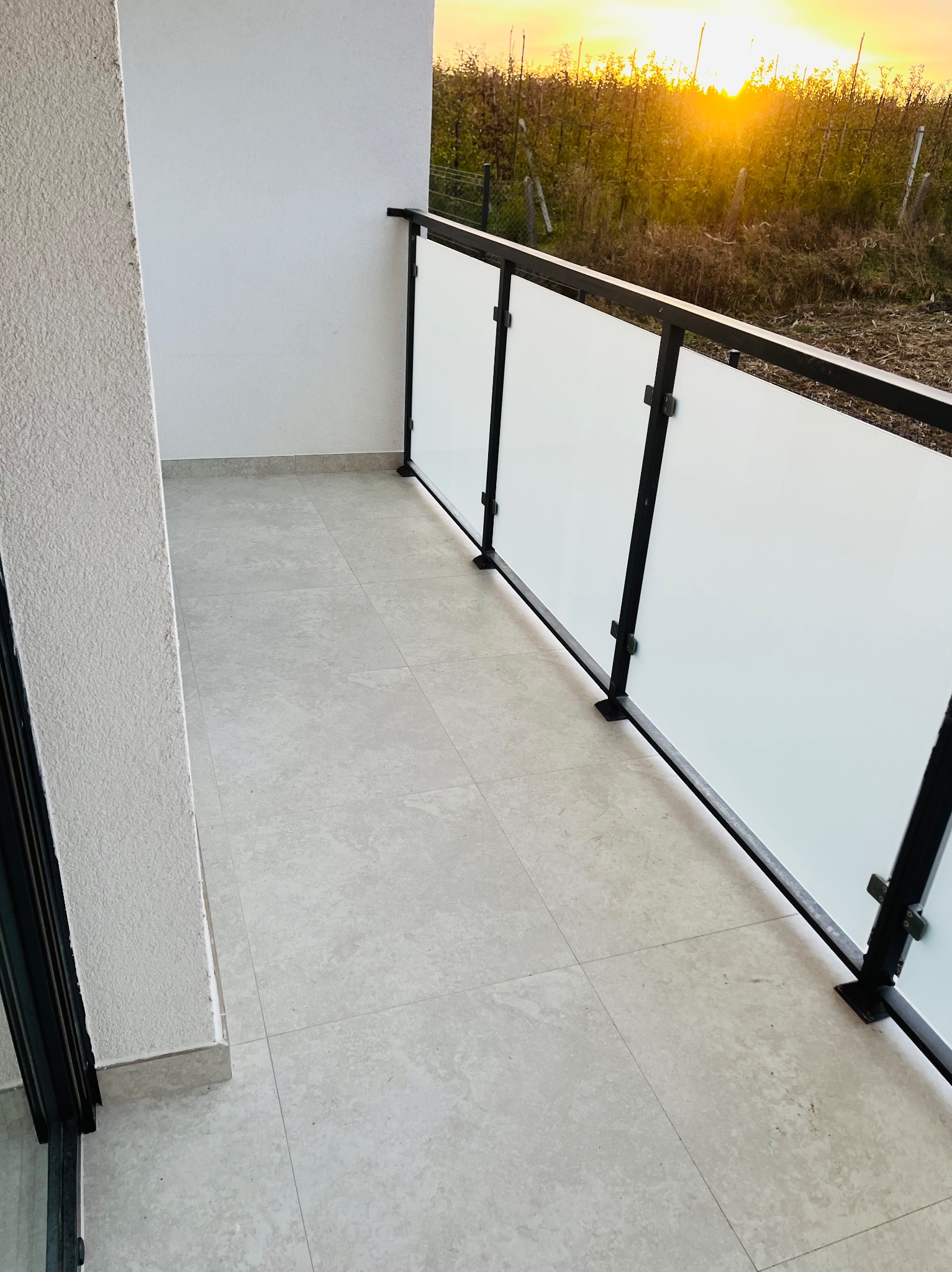 Wykończony balkon z jasnymi płytkami podłogowymi i nowoczesną balustradą z czarną ramą i mlecznym szkłem, widok na zachodzące słońce za drzewami.