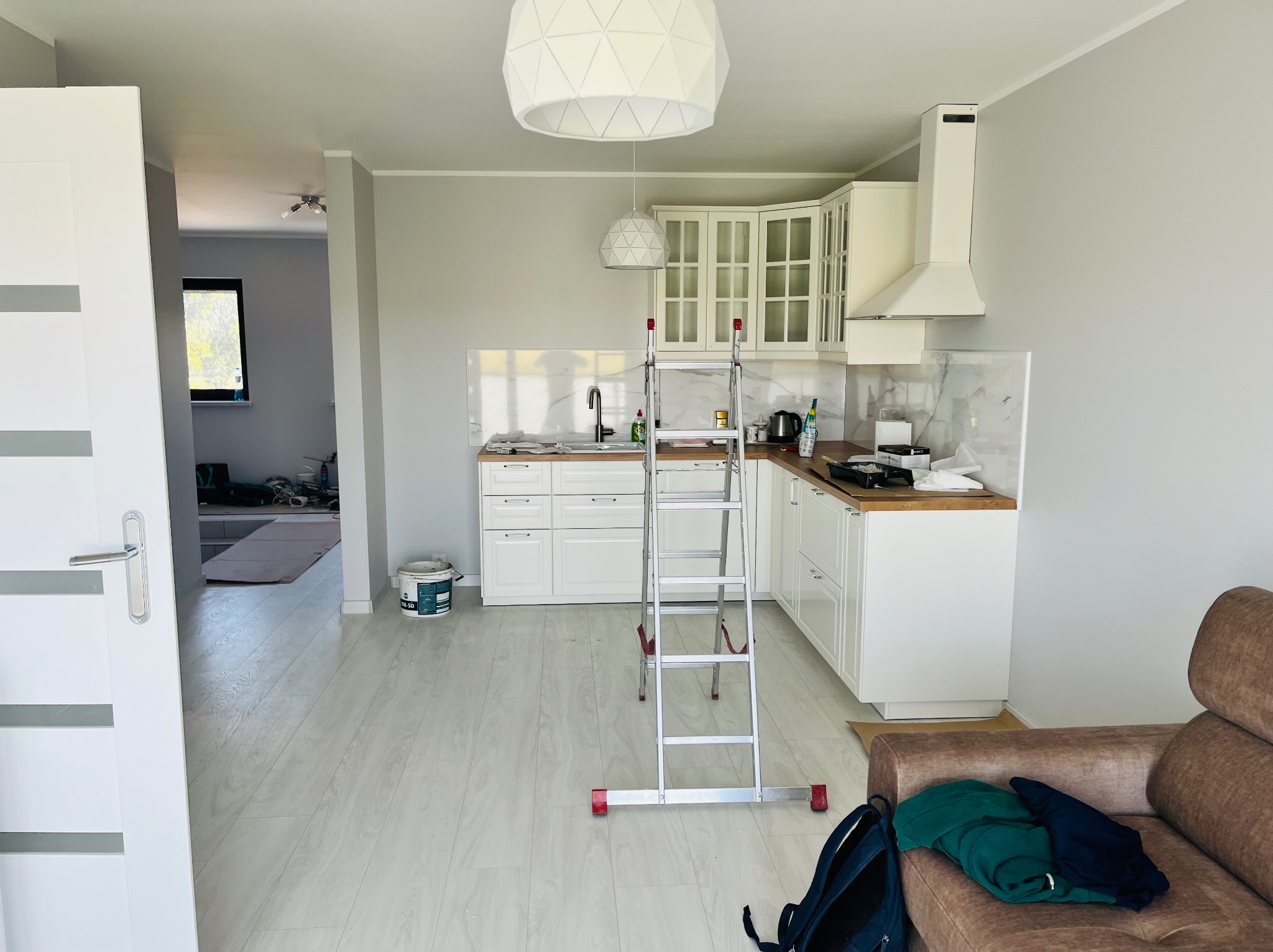 Wnętrze jasnej kuchni w trakcie prac wykończeniowych z rozstawioną drabiną, widoczne białe meble kuchenne, podłoga z paneli, ściany w odcieniach szarości oraz wiadro z farbą w tle.