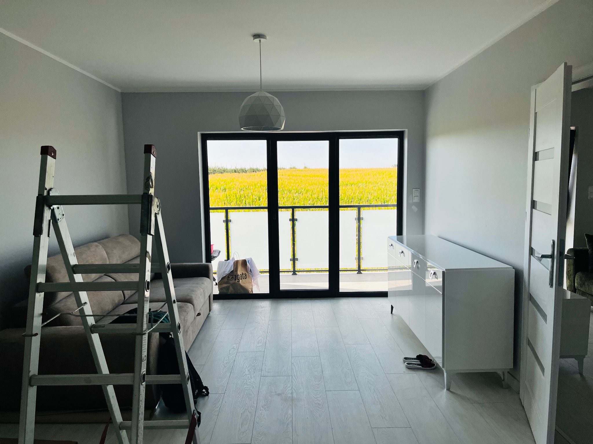 Jasne wnętrze pokoju po odświeżeniu, z drabiną malarską, szarymi ścianami, białymi meblami i dużym oknem balkonowym z widokiem na pole.