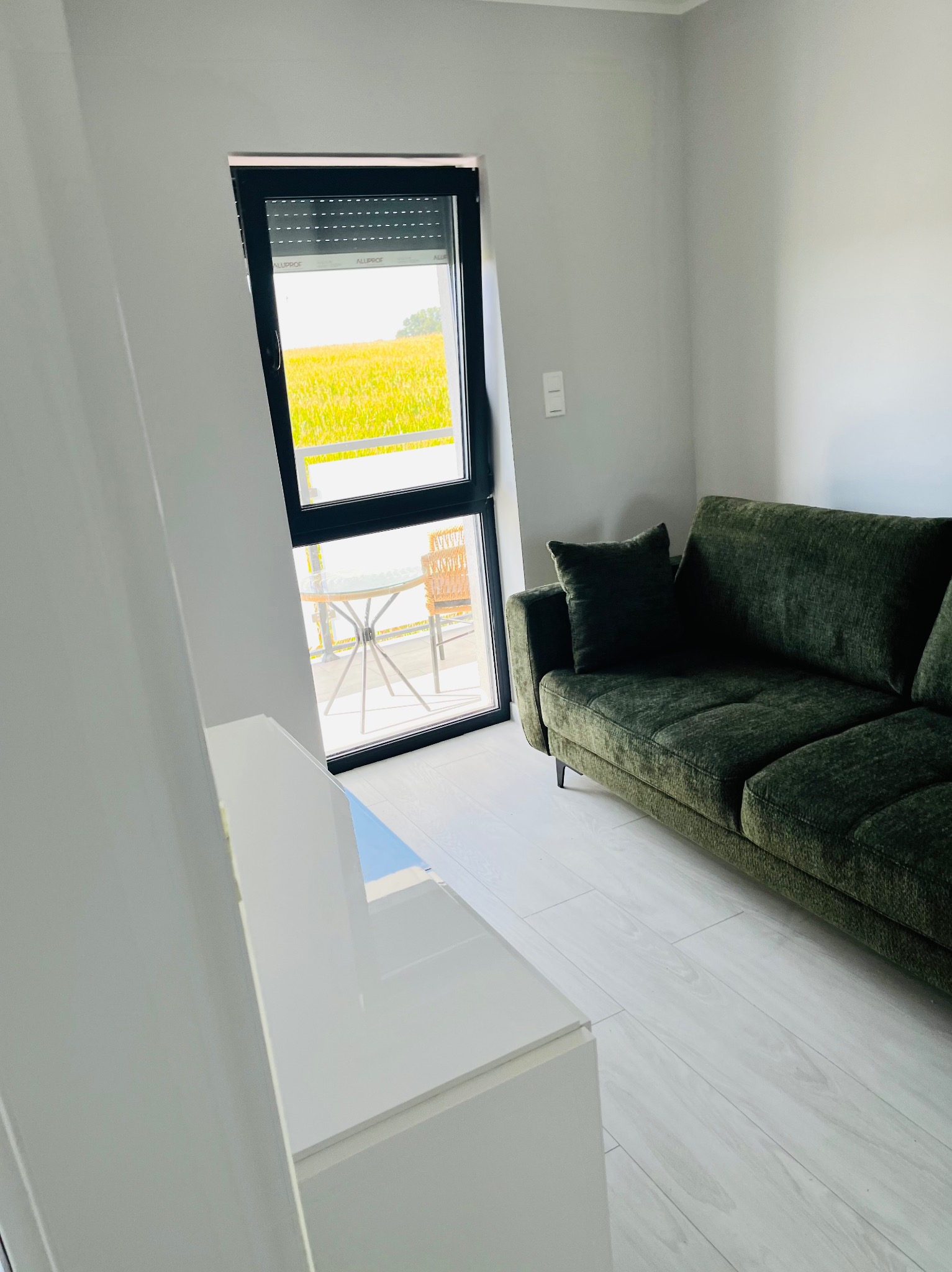 Nowoczesny salon z zieloną sofą i widokiem na pole z balkonu, jasne ściany i białe panele podłogowe, minimalistyczny wystrój.