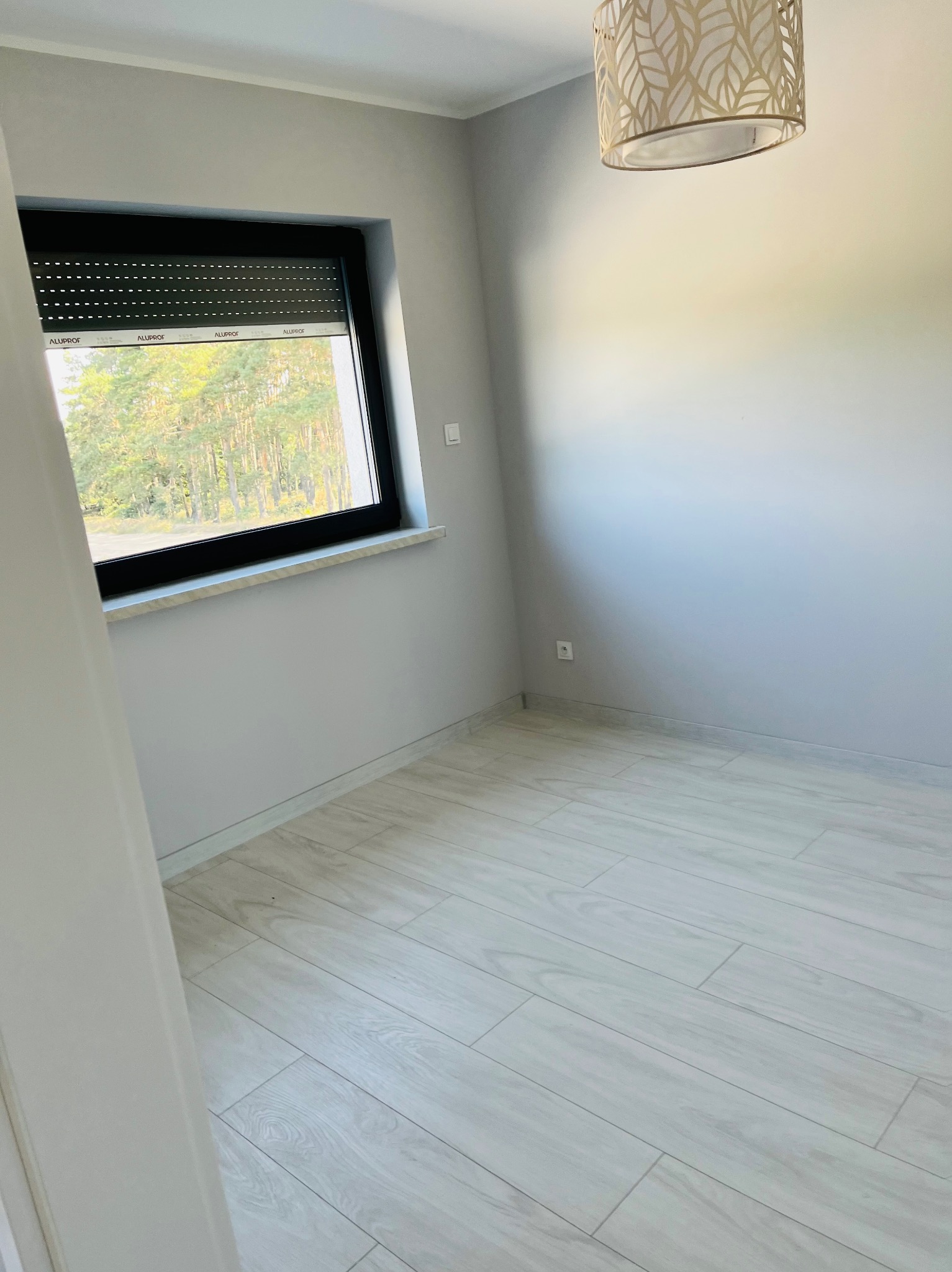 Pusty pokój z szarymi ścianami, jasną podłogą imitującą drewno i roletami w oknie, widok na drzewa z zewnątrz, designerska lampa sufitowa.