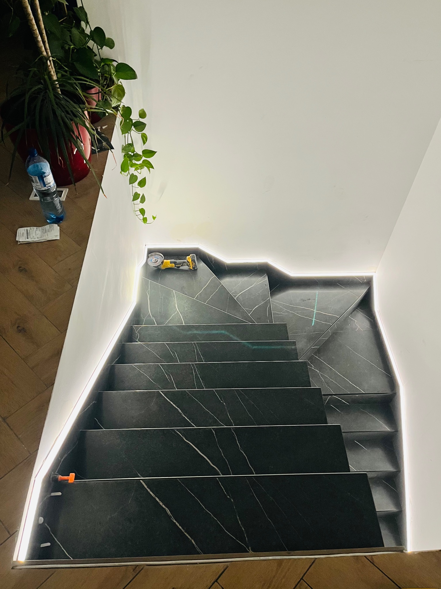 Schody wyłożone czarnym gresem z białymi żyłami, podświetlone listwą LED, z narzędziem do cięcia płytek na górnym stopniu, widok z góry.