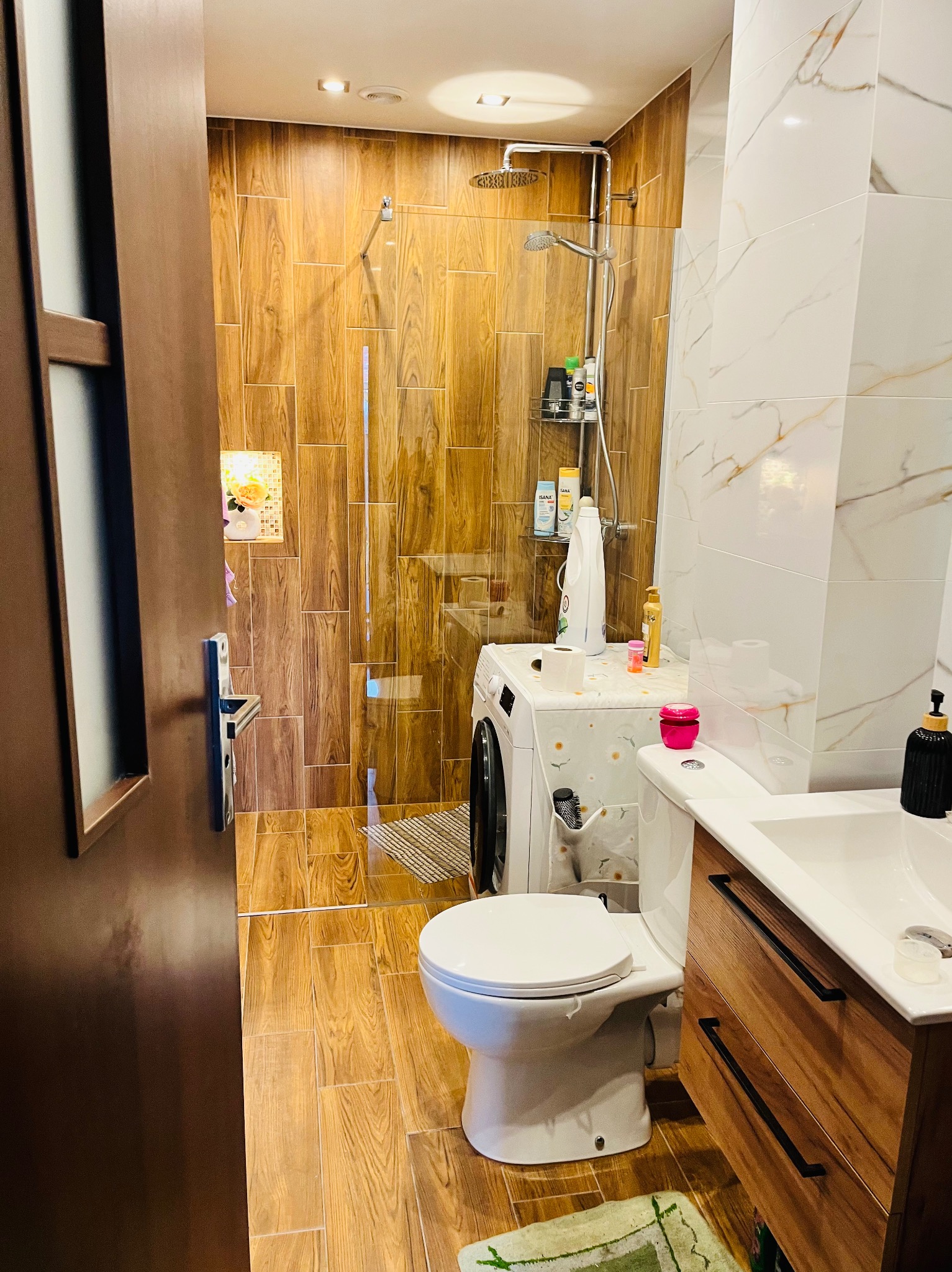 Nowoczesna łazienka z drewnopodobnymi płytkami, kabiną prysznicową typu walk-in i pralką. Umywalka z drewnianą szafką, WC kompakt. Jasne, ciepłe oświetlenie.