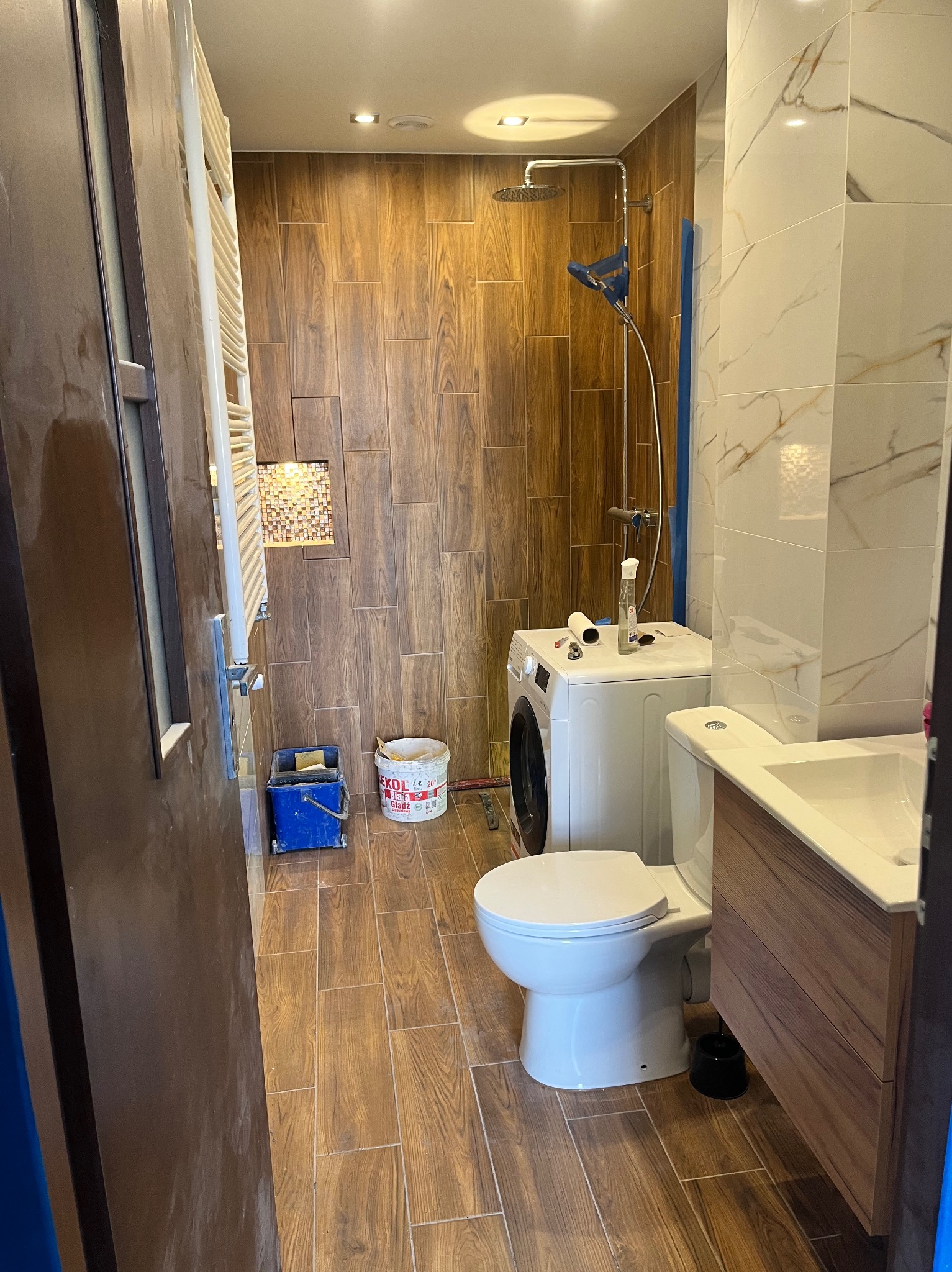 Wykończona łazienka z drewnopodobnymi płytkami, kabiną prysznicową typu walk-in, umywalką z szafką. Widoczna pralka i akcesoria malarskie.