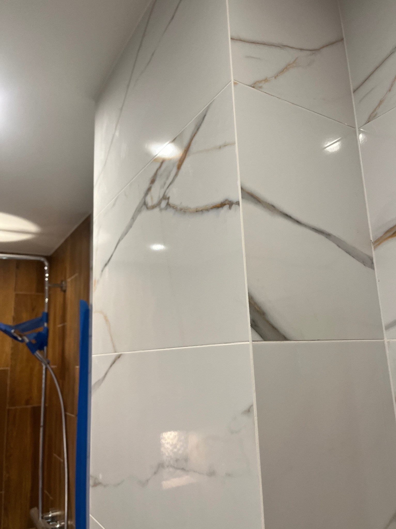 Białe, błyszczące płytki imitujące marmur z brązowymi żyłkami, ułożone na ścianie w łazience. Widoczny fragment kabiny prysznicowej z niebieskim profilem.