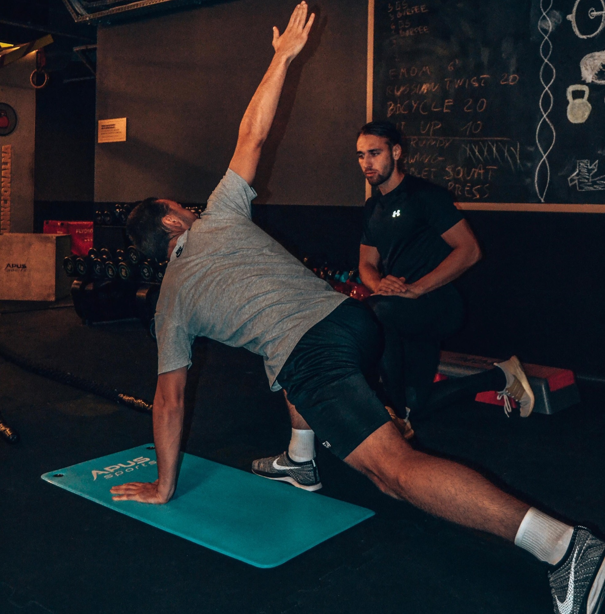 Mężczyzna wykonuje ćwiczenie rozciągające na macie w siłowni, pod okiem trenera. Na tablicy wypisane nazwy ćwiczeń i liczby powtórzeń.