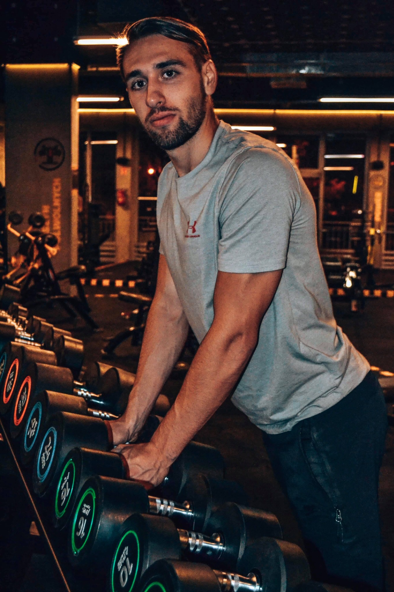 Młody mężczyzna w szarej koszulce opiera się o stojak z hantlami w siłowni z ciepłym oświetleniem, spoglądając w obiektyw.