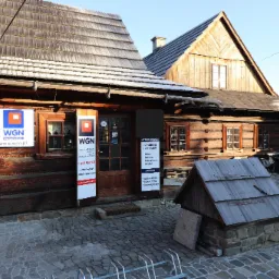 Tradycyjny drewniany budynek z szyldami agencji nieruchomości WGN, oferującej sprzedaż i wynajem, z małą drewnianą studzienką na pierwszym planie.