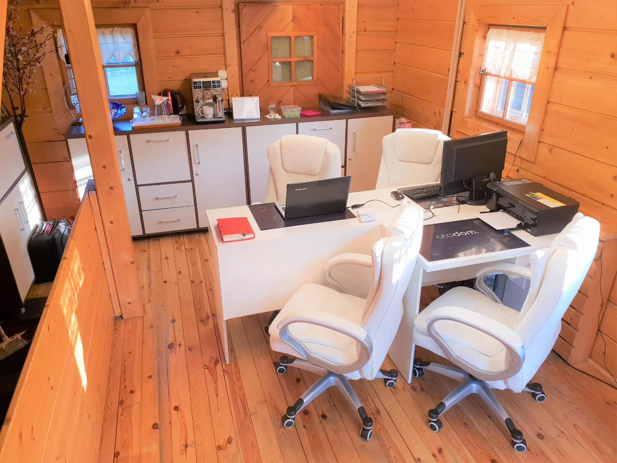 Wnętrze biura w drewnianym domku, z trzema białymi krzesłami obrotowymi, białym biurkiem z laptopem, monitorem, drukarką i akcesoriami biurowymi, z drewnianą podłogą i ścianami z desek.