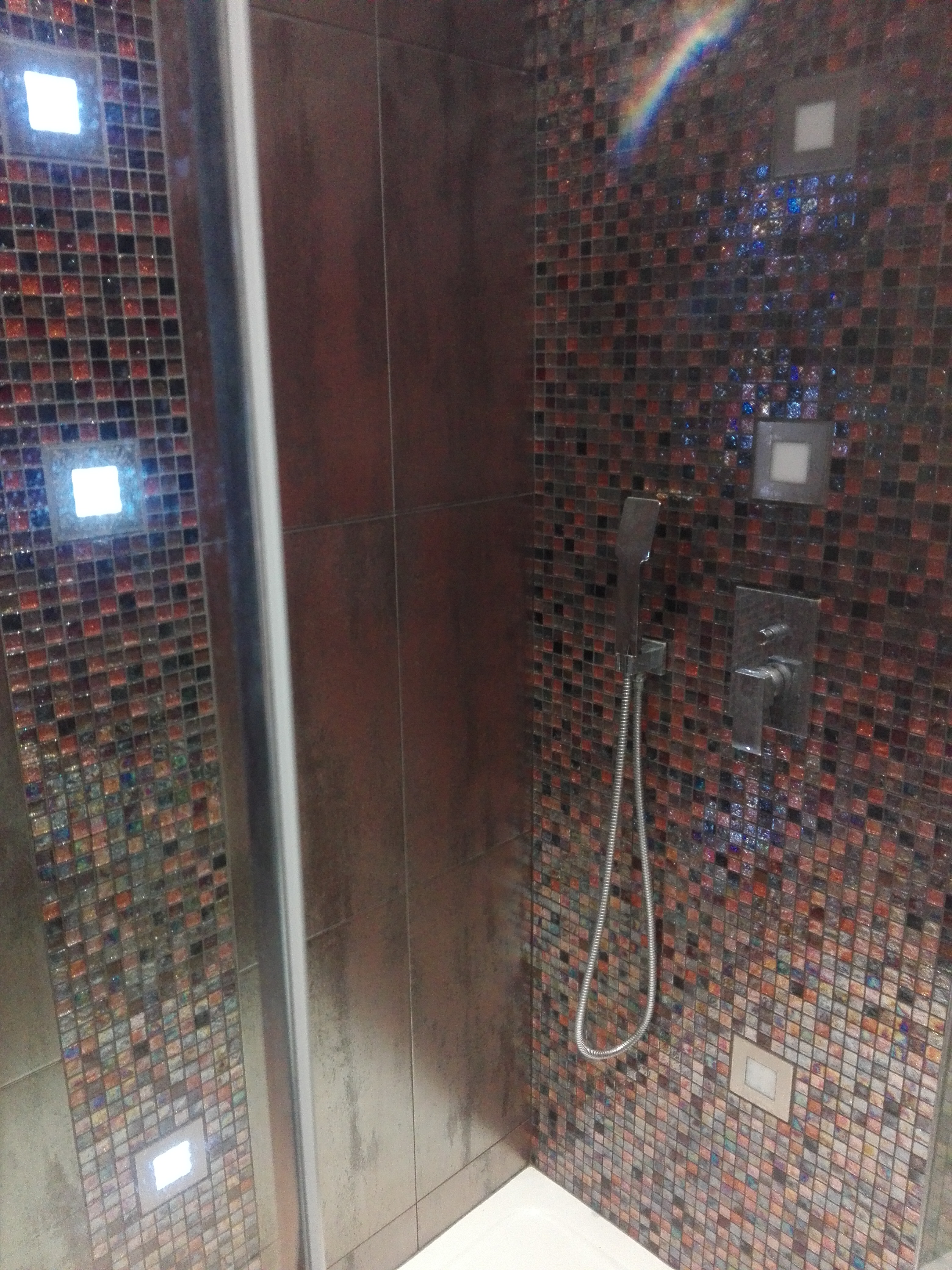 Kabina prysznicowa z mozaiką w odcieniach czerwieni, brązu i czerni, wbudowane oświetlenie punktowe, bateria i słuchawka prysznicowa w kolorze srebrnym.