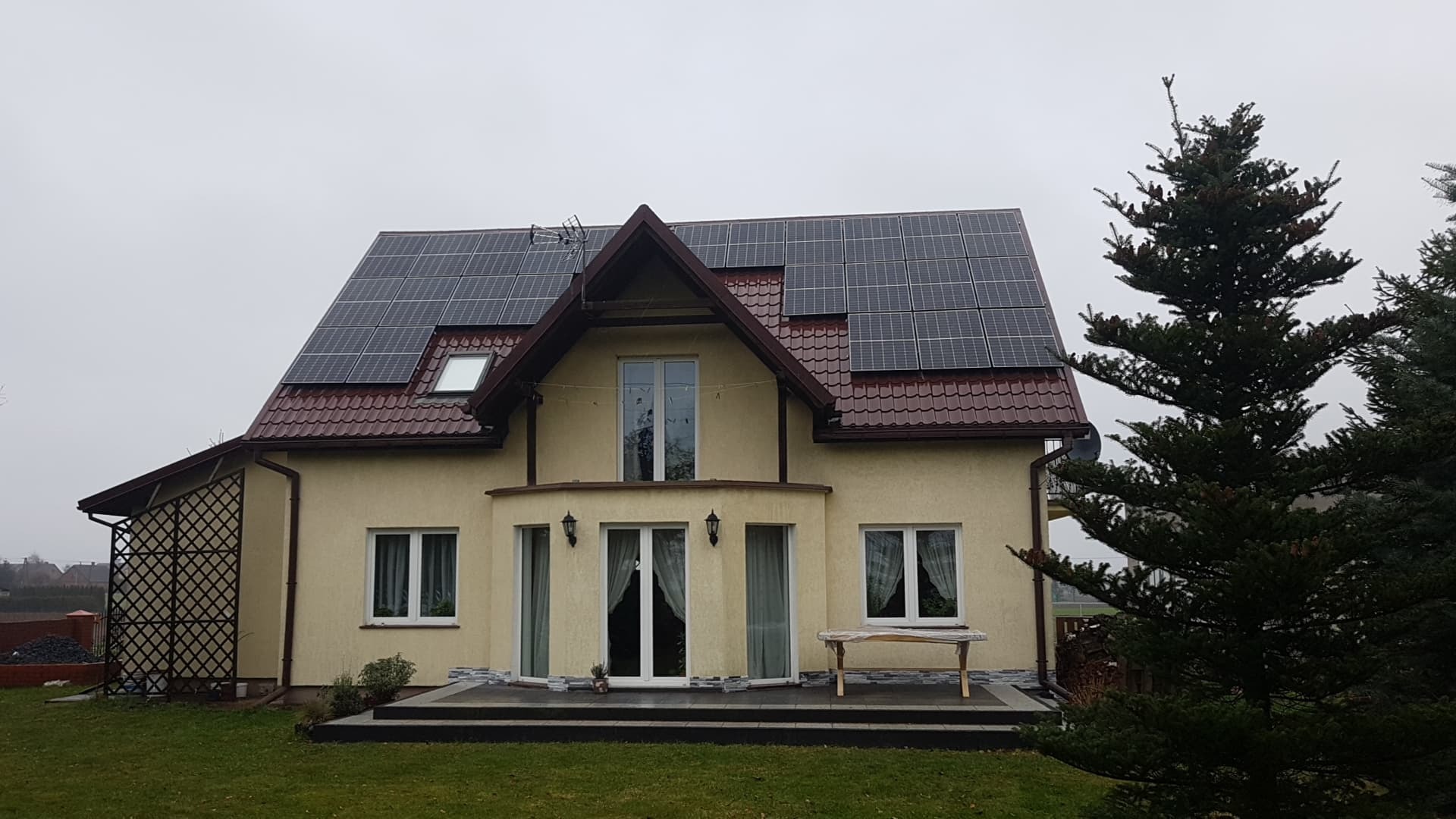 Żółty dom jednorodzinny z panelami słonecznymi na dachu z czerwonej dachówki, widok z przodu w pochmurny dzień.