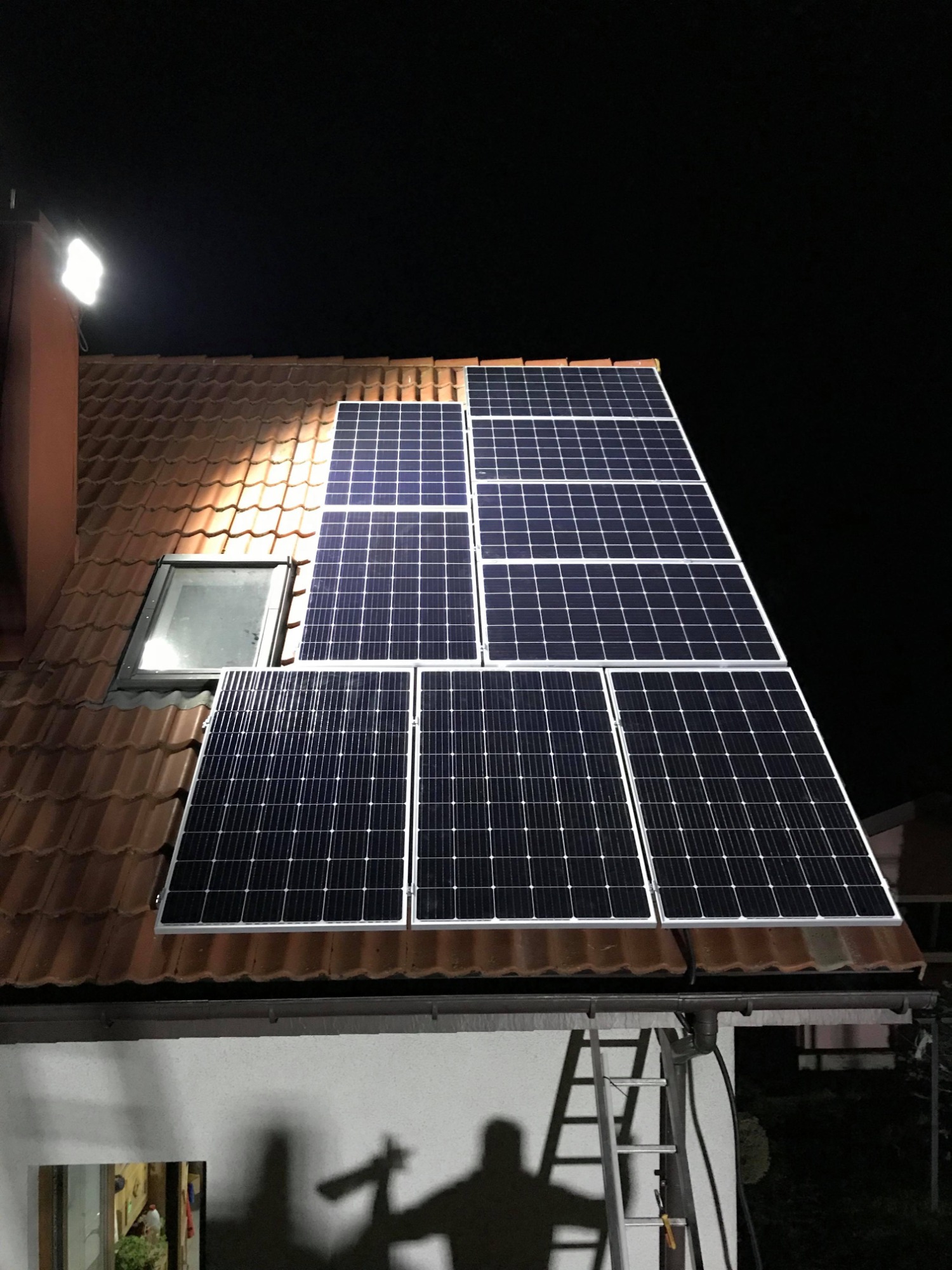 Instalacja paneli słonecznych na dachu domu jednorodzinnego oświetlona reflektorem, widoczna drabina i cień osoby.
