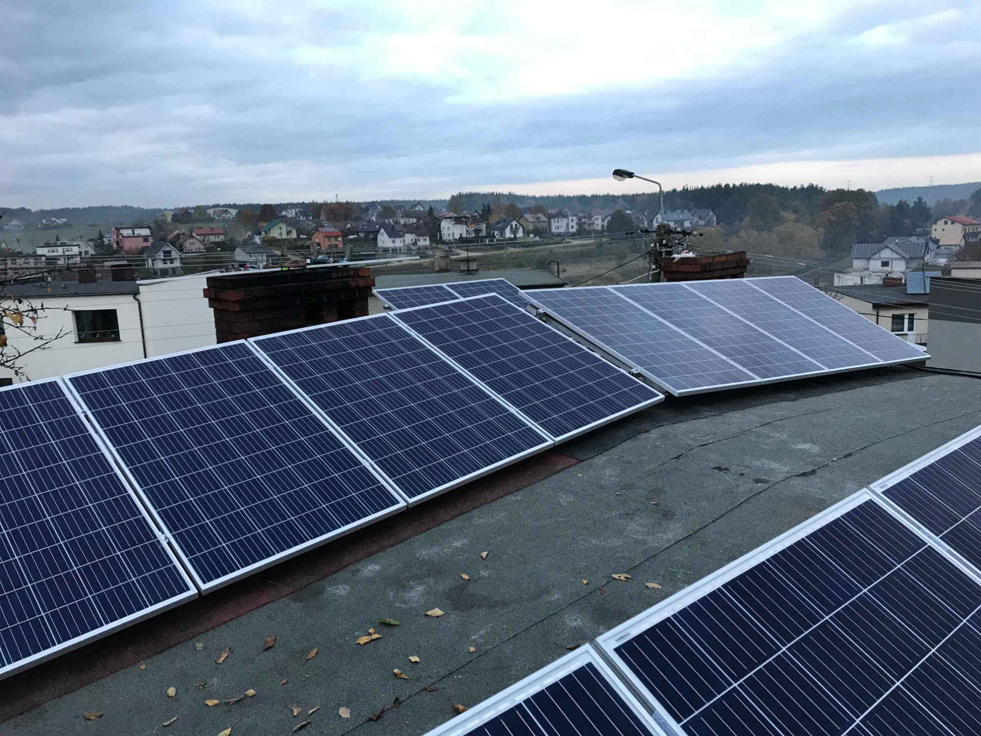 Instalacja fotowoltaiczna na płaskim dachu budynku mieszkalnego, widok na miasto w tle, pochmurne niebo.