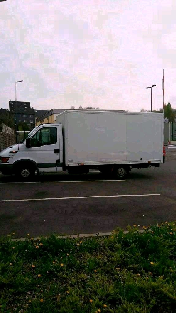 Biała furgonetka dostawcza zaparkowana na parkingu, widok z boku, zieleń i budynki w tle.