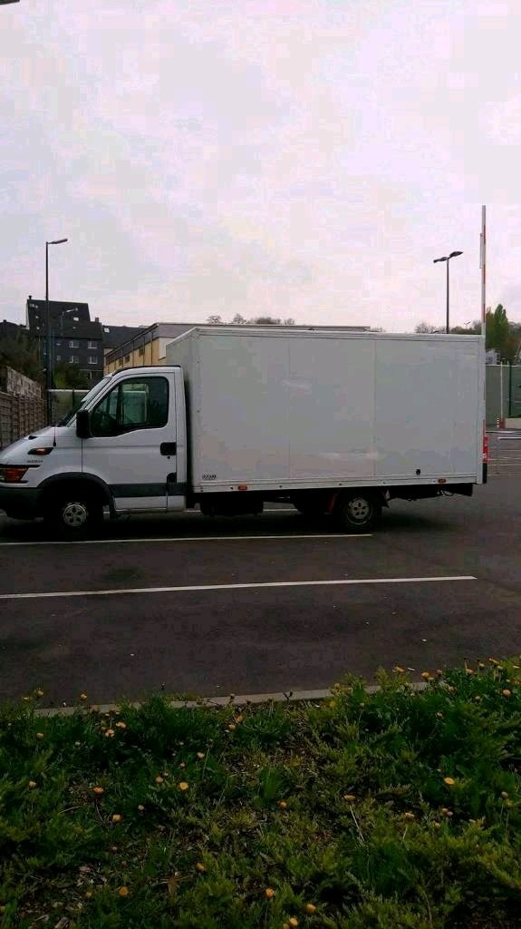 Biała furgonetka dostawcza zaparkowana na parkingu, z widocznymi budynkami w tle i krzewami z żółtymi kwiatami na pierwszym planie.