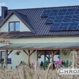 Dom jednorodzinny z brązowym dachem i panelami słonecznymi, widok zza pola zboża; biały van z otwartymi drzwiami zaparkowany obok.