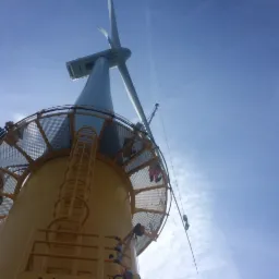 Żółta wieża turbiny wiatrowej z drabinką i platformą, widok z dołu na tle błękitnego nieba, liny i osoby w kombinezonach na platformie.