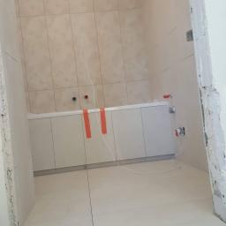 MGL Ochir-Erdene Tserensodnom - Łazienka w trakcie remontu: świeżo położone, beżowe płytki na ścianach i podłodze, widoczne instalacje wodne i elektryczne, zabezpieczona wanna oraz narzędzia glazurnicze w rogu pomieszczenia.