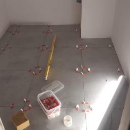 MGL Ochir-Erdene Tserensodnom - Szare płytki podłogowe w trakcie układania, widoczne poziomica, pojemnik z klipsami poziomującymi i gąbka do czyszczenia fug.