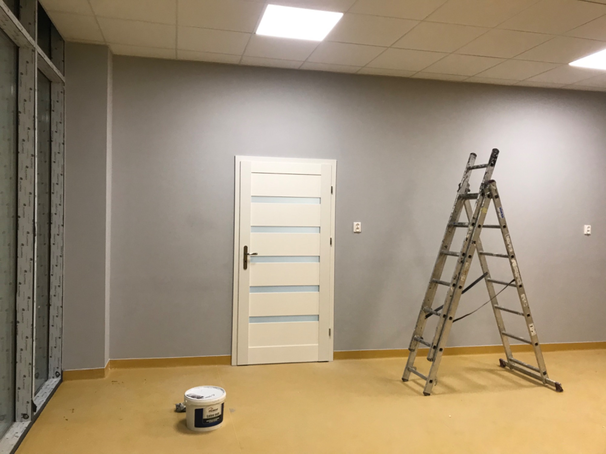 Szare ściany w trakcie odświeżania, widoczna drabina, puszka z farbą i białe drzwi z poziomymi przeszkleniami w pomieszczeniu biurowym przy ul. Rotmistrza Pileckiego 67 w Warszawie.