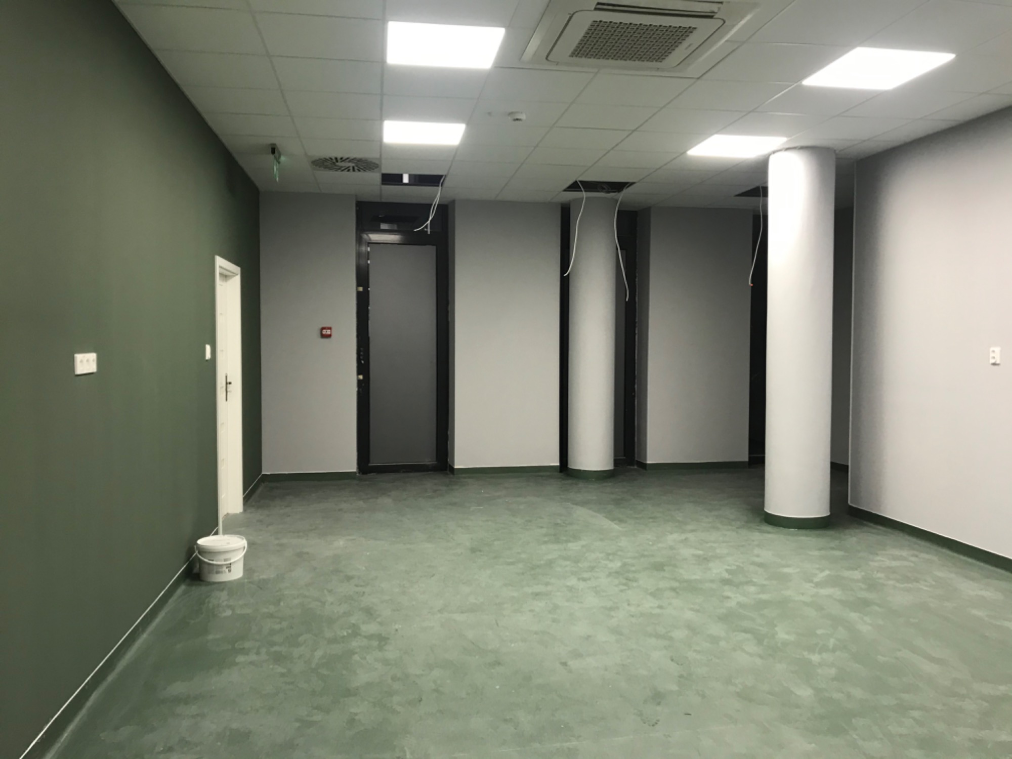 Wnętrze biurowe w trakcie malowania: oliwkowa ściana, betonowa posadzka w odcieniach zieleni, białe kolumny i sufit z kasetonami, widoczne kable zwisające z sufitu i wiadro z farbą przy ścianie.