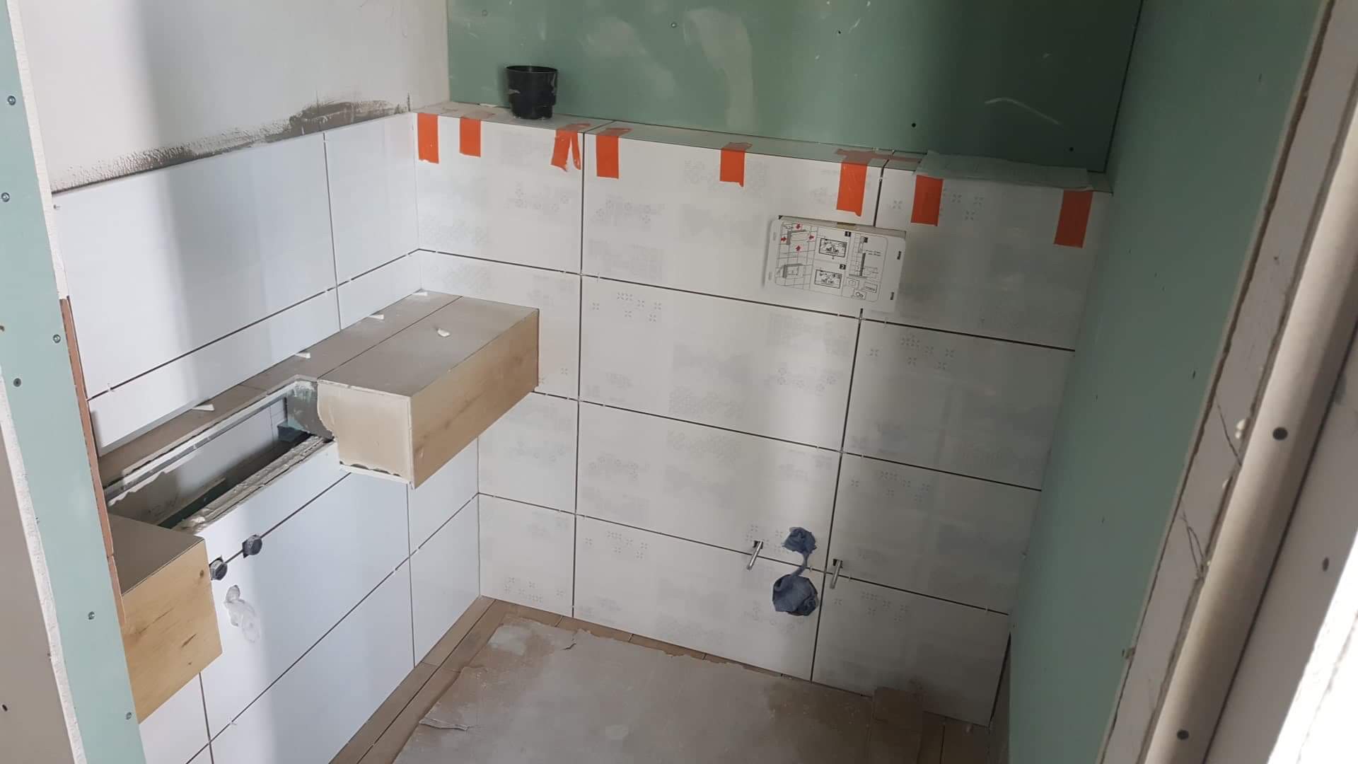 Łazienka w trakcie remontu w Warszawie: ściany wyłożone białymi płytkami, częściowo zabezpieczone pomarańczową taśmą, widoczne fragmenty instalacji oraz płyty gipsowo-kartonowe na ścianach.