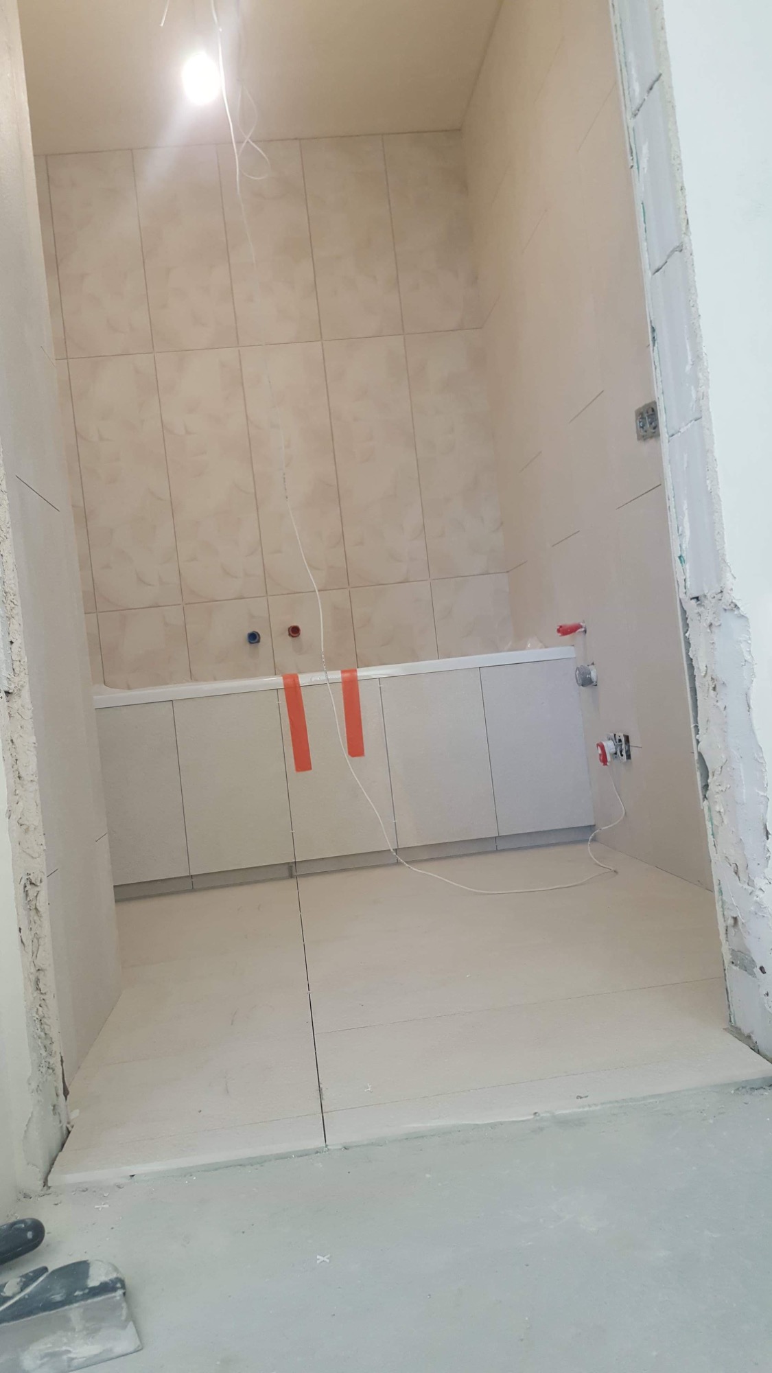 Łazienka w trakcie remontu: świeżo położone, beżowe płytki na ścianach i podłodze, widoczne instalacje wodne i elektryczne, zabezpieczona wanna oraz narzędzia glazurnicze w rogu pomieszczenia.