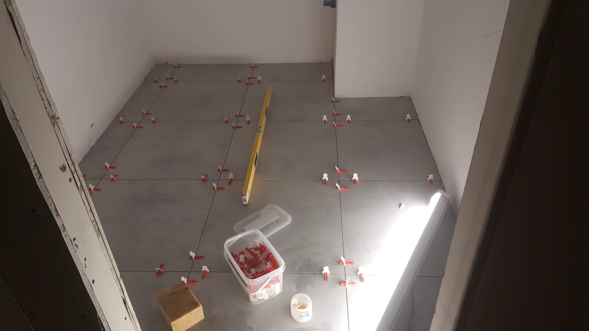 Szare płytki podłogowe w trakcie układania, widoczne poziomica, pojemnik z klipsami poziomującymi i gąbka do czyszczenia fug.