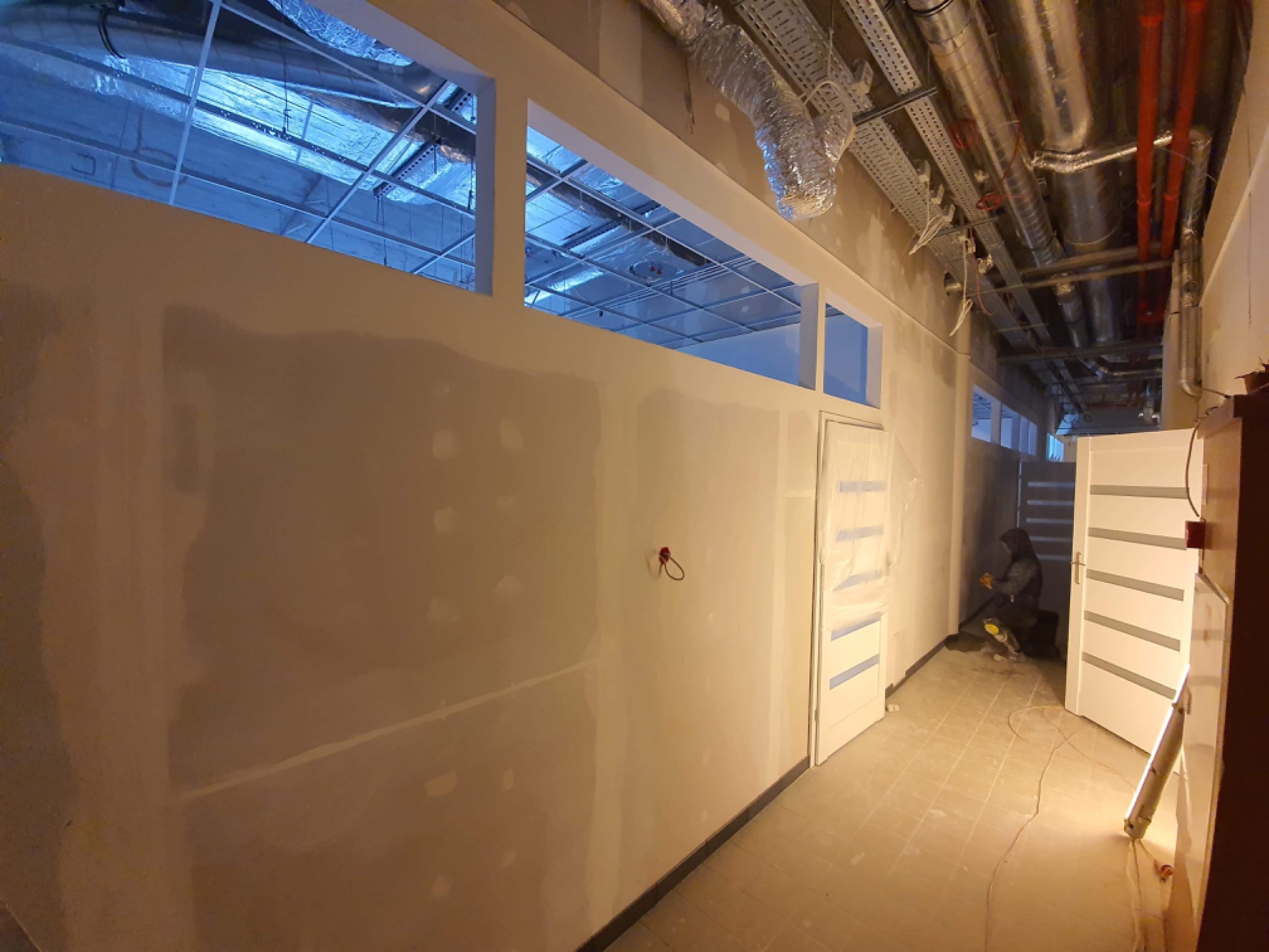 Korytarz biurowy w trakcie prac wykończeniowych; ściany przygotowane do malowania, widoczne szpachlowane fragmenty, drzwi z folią ochronną, sufit z instalacjami wentylacyjnymi, pracownik w tle...