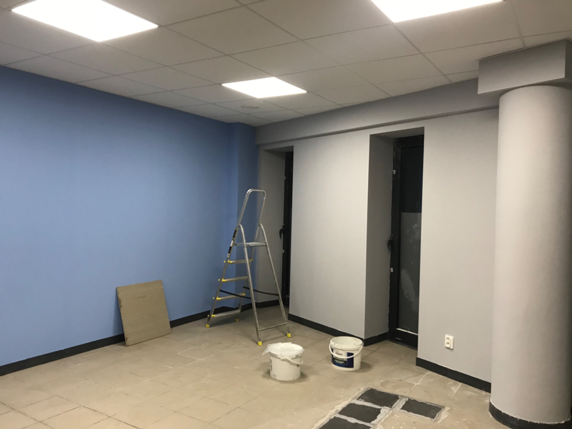 Wnętrze biurowe w trakcie malowania: jedna ściana na niebiesko, reszta na szaro, drabina, dwa wiadra z farbą, fragment podłogi z odkrytymi płytkami.