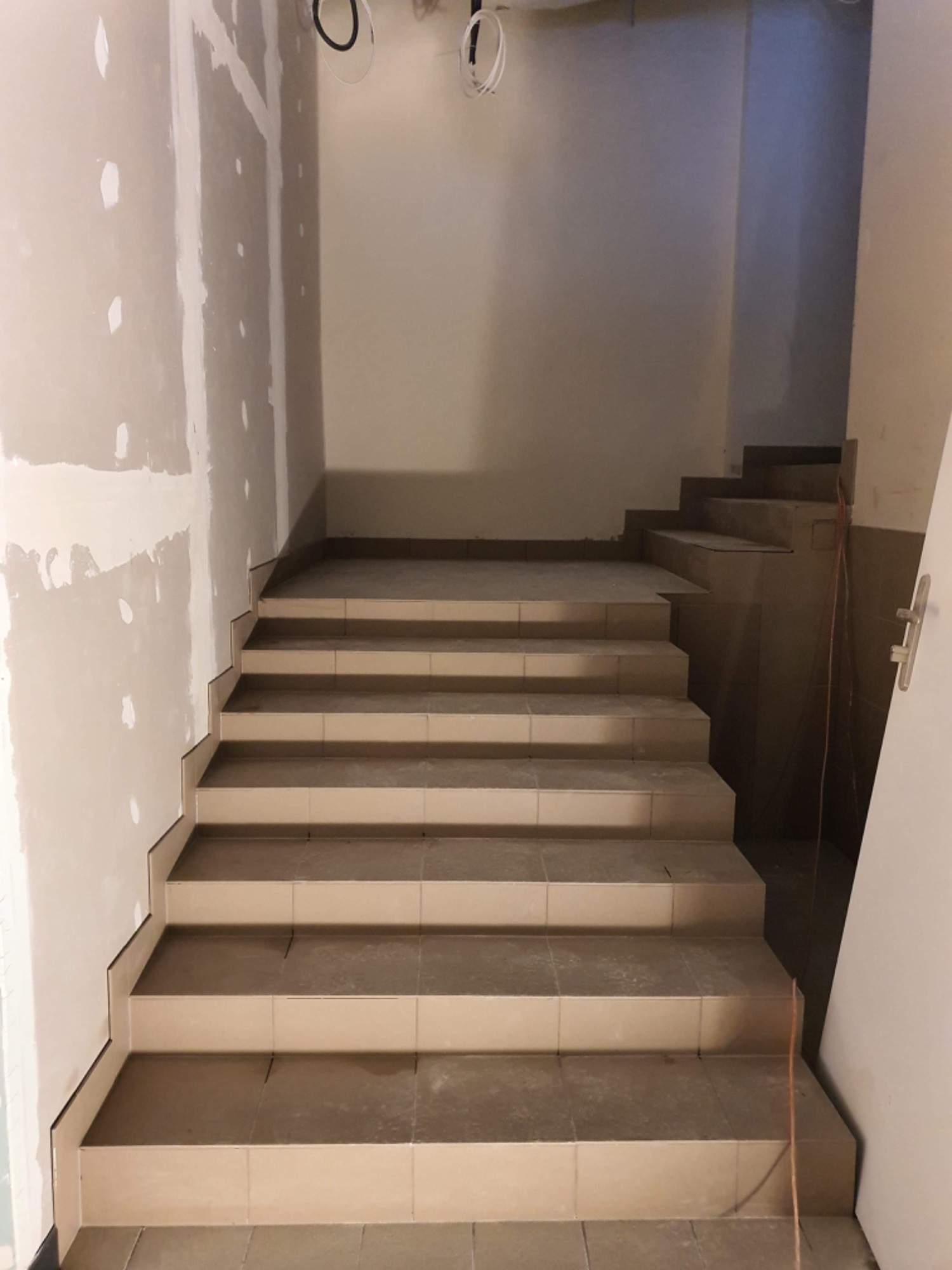 Klatka schodowa w trakcie wykańczania: ułożone płytki na schodach, ściany częściowo wyszpachlowane, widoczne przewody na suficie.