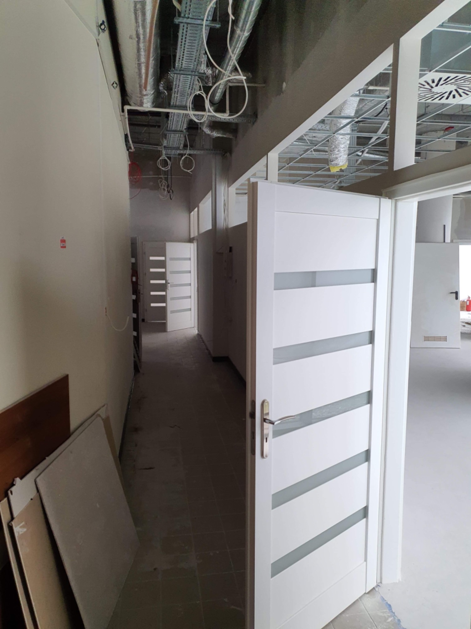 Wnętrze biura w trakcie remontu, widoczne otwarte białe drzwi z poziomymi przeszkleniami, instalacje pod sufitem i płyty gipsowe oparte o ścianę.
