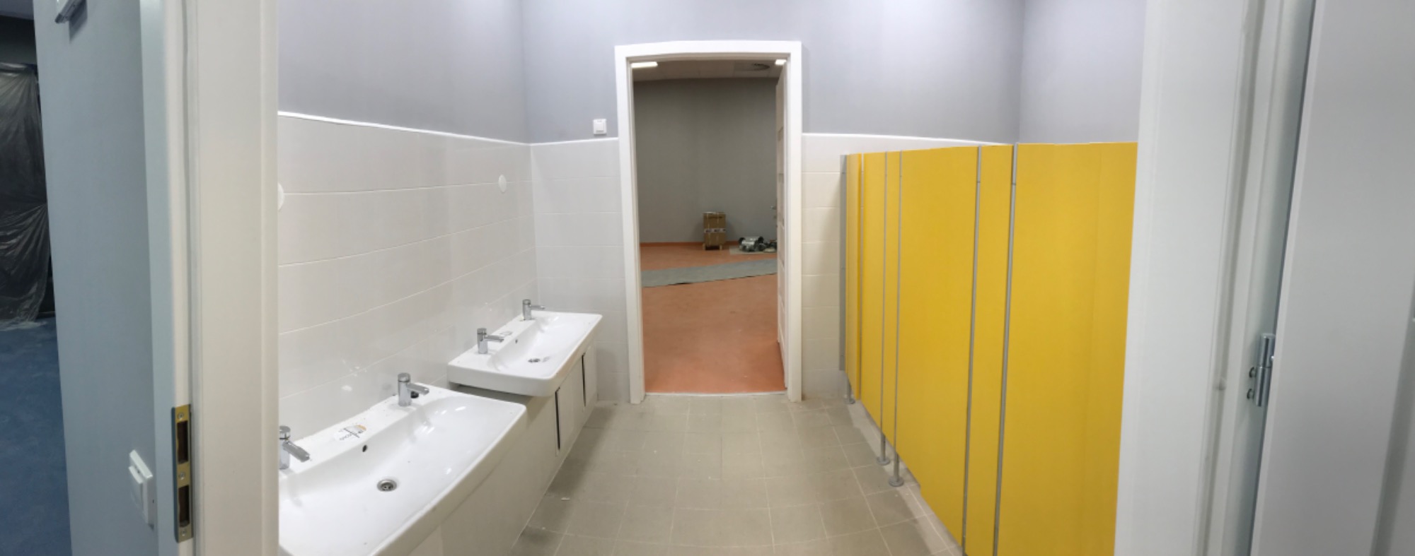 Wnętrze publicznej toalety z dwoma umywalkami z szafkami, białymi płytkami na ścianach, szarą górną częścią ścian, żółtymi kabinami WC i widocznym wejściem do pomieszczenia obok.