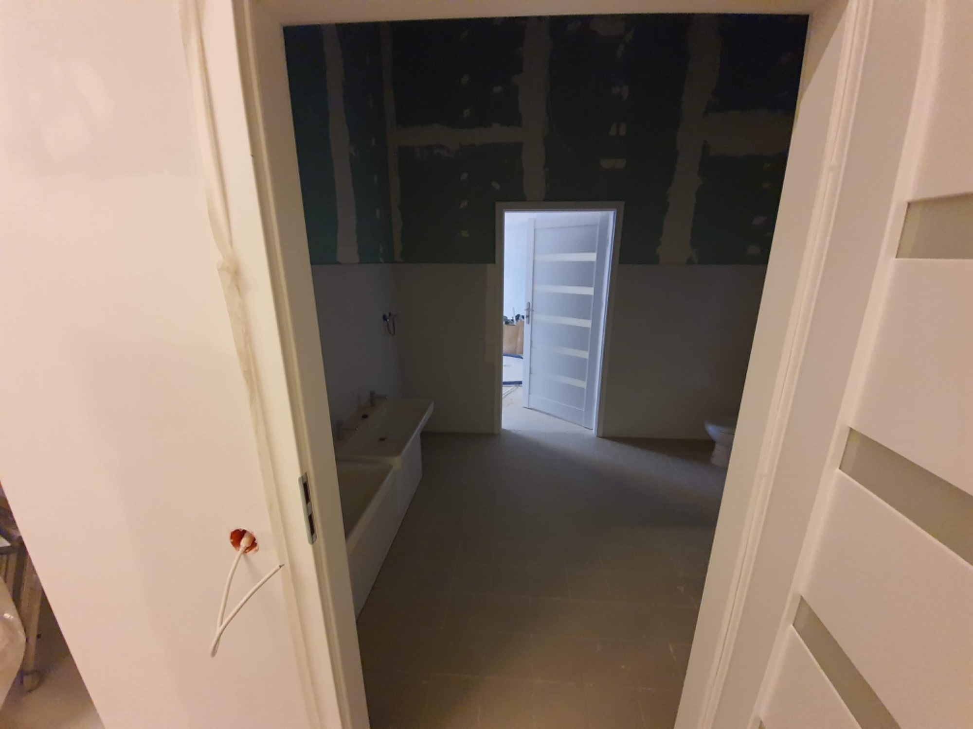 Łazienka w trakcie remontu: widoczne płyty kartonowo-gipsowe na ścianach, surowa instalacja elektryczna wystająca ze ściany obok ościeżnicy, oraz umywalka w głębi pomieszczenia.