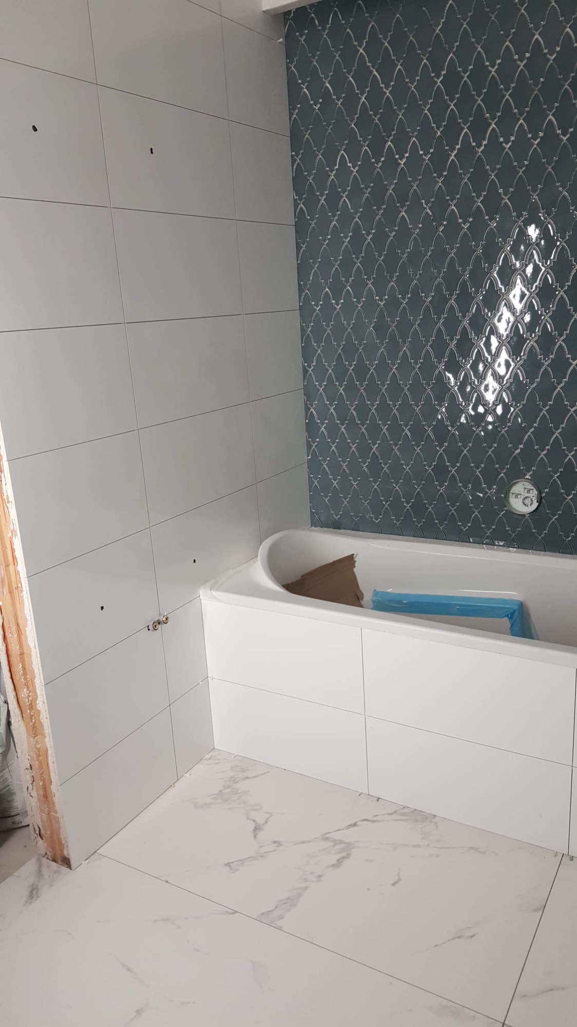 Łazienka w trakcie wykańczania: białe płytki ścienne i podłogowe z marmurowym wzorem, dekoracyjna ściana z ciemnych płytek o wzorze arabeski, zabudowana wanna z elementami zabezpieczającymi.
